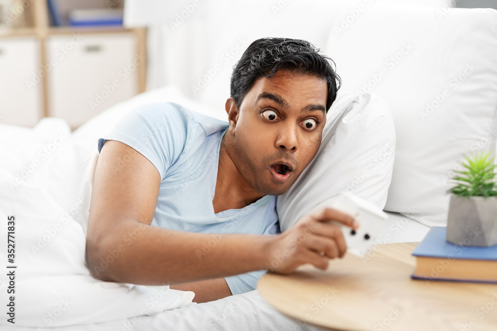 people, bedtime and rest concept - overslept indian man looking at alarm clock lying in bed at home people, bedtime and rest concept - overslept indian man looking at alarm clock lying in bed at home