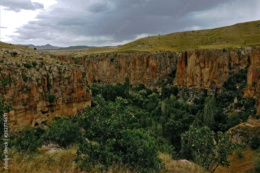 Ihlara Canyon in Cappadocia. The grass-covered plain is divided into parts by a gorge with steep rocky slopes. At the bottom, trees grow. Cloudy. In the distance are the silhouettes of mountains.