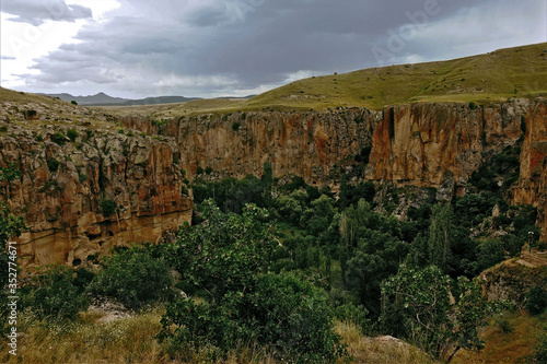 Ihlara Canyon in Cappadocia. The grass-covered plain is divided into parts by a gorge with steep rocky slopes. At the bottom  trees grow. Cloudy. In the distance are the silhouettes of mountains.