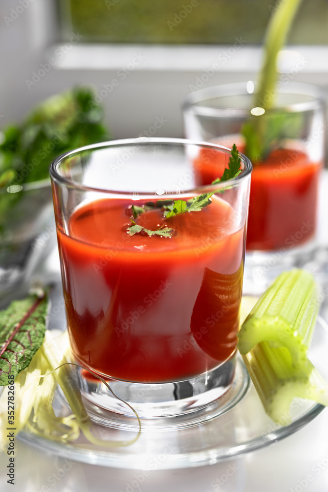 Two glasses of tomato juice with parsley and celery decorations, stand near the window, morning sunlight shines, shallow depth of field, selective focus. Natural drinks concept. Two glasses of tomato juice with parsley and celery decorations, stand near the window, morning sunlight shines, shallow depth of field, selective focus. Natural drinks concept.