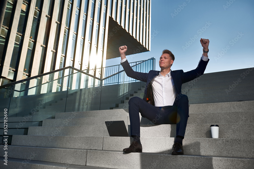 Young businessman sits on the stairs between high-rise buildings, celebrates his success and puts his arms up 