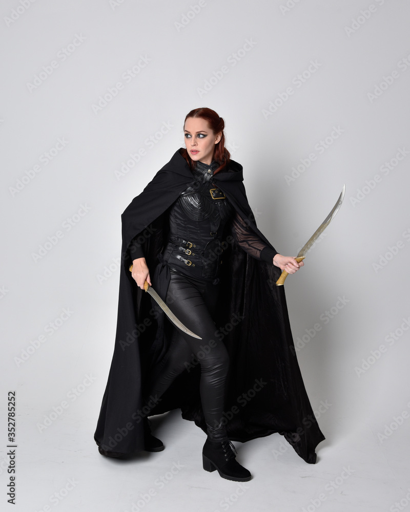 fantasy portrait of a woman with red hair wearing dark leather assassin costume with long black cloak. Full length standing pose holding a sword isolated against a studio background. fantasy portrait of a woman with red hair wearing dark leather assassin costume with long black cloak. Full length standing pose holding a sword isolated against a studio background.