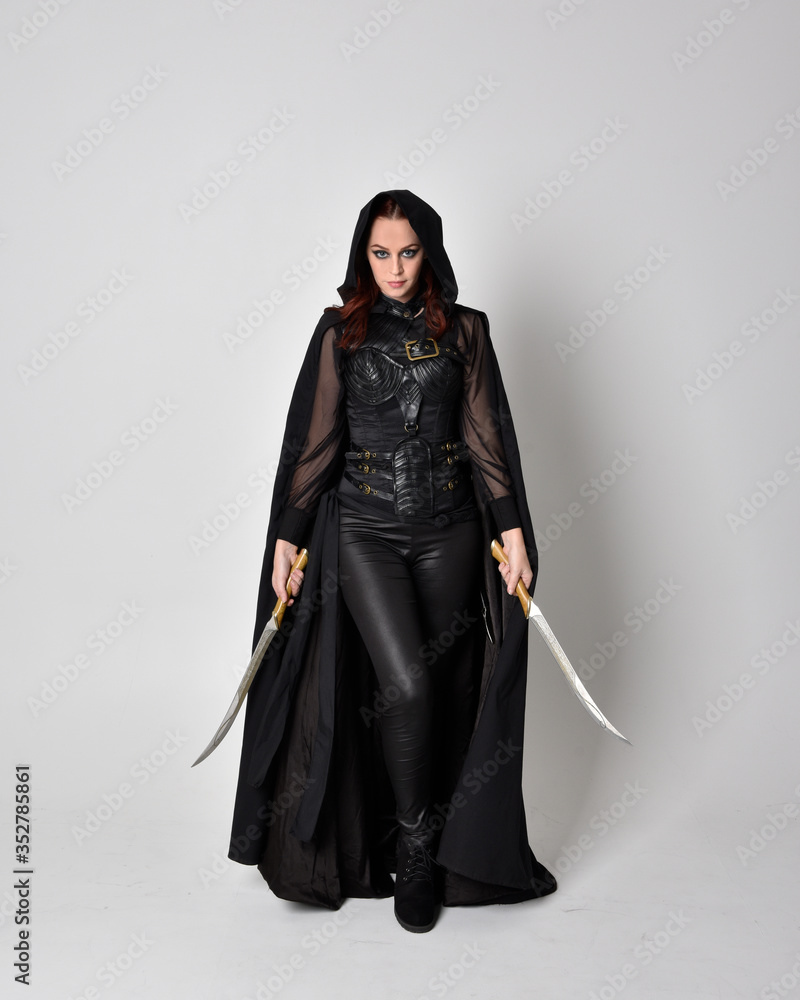 fantasy portrait of a woman with red hair wearing dark leather assassin costume with long black cloak. Full length standing pose holding a sword isolated against a studio background.