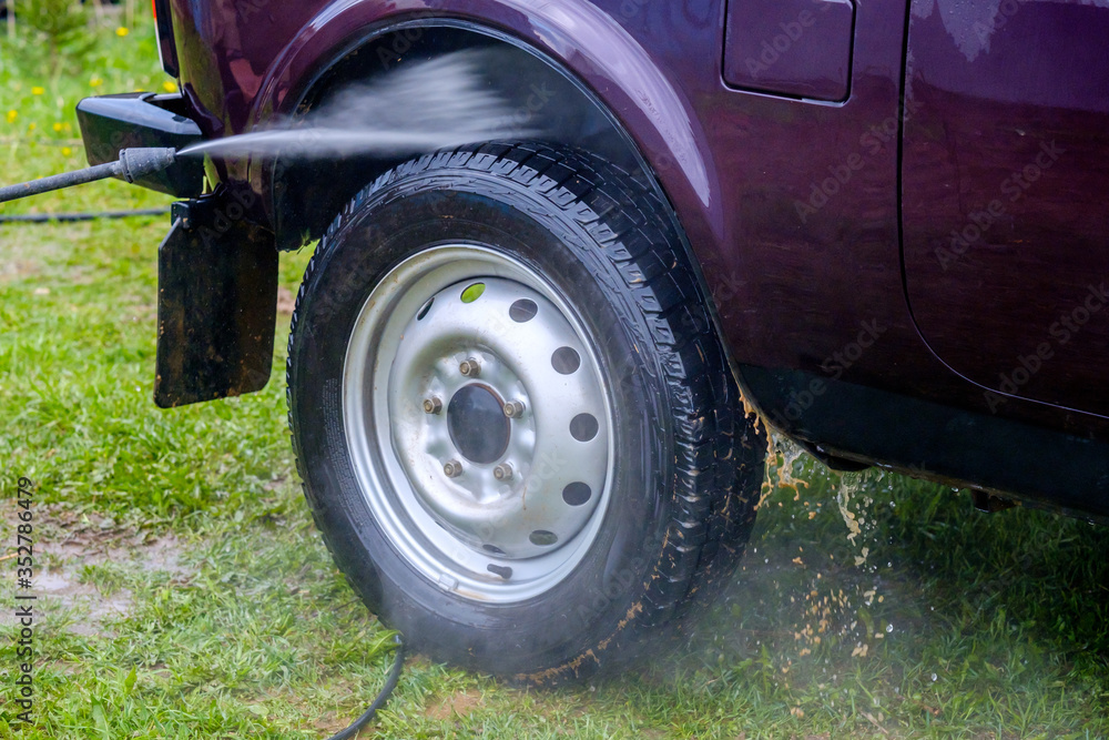 Car wash in the afternoon outdoors with a high-pressure apparatus. A powerful spray with water splashes away the dirt from the purple body of the car, from glass, wheels and tires.
