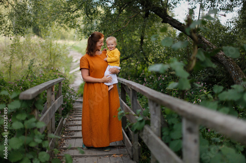 Caucasian mother with baby in her arms in a park on a wooden bridge  mother in a linen dress holds a one-year-old baby in her arms  walking in the park  concept motherhood and summer  walks near home