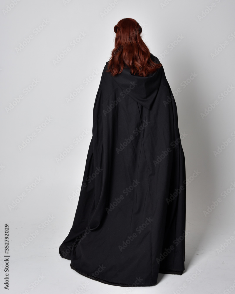 fantasy portrait of a woman wearing long black cloak. Full length standing pose  with back to the camera, isolated against a studio background.