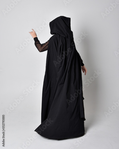 fantasy portrait of a woman wearing long black cloak. Full length standing pose with back to the camera, isolated against a studio background.