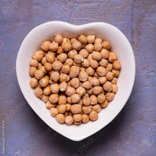 view from top. Dried chickpeas in the foreground in a white ceramic bowl in the shape of a heart on a textured background