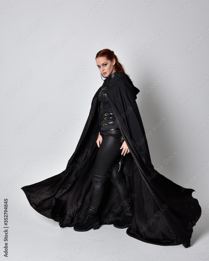 fantasy portrait of a woman with red hair wearing dark leather assassin  costume with long black cloak. Full length standing pose in side profile  isolated against a studio background. Stock Photo