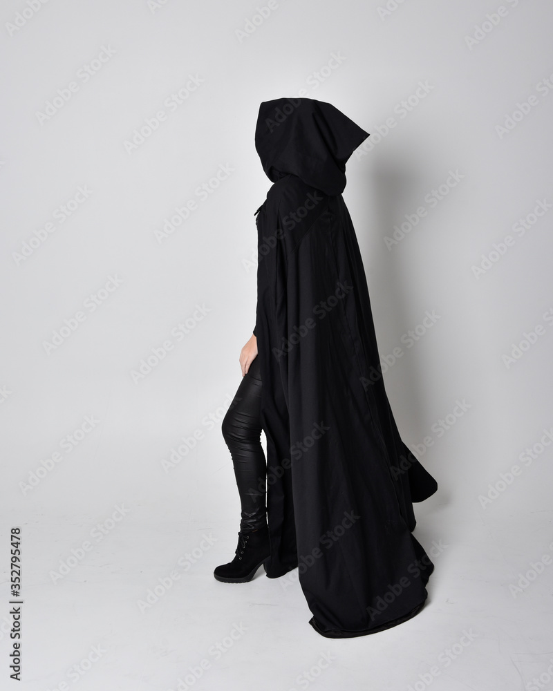fantasy portrait of a woman with red hair wearing dark leather assassin costume with long black cloak. Full length standing pose in side profile isolated against a studio background.