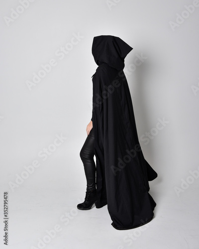 fantasy portrait of a woman with red hair wearing dark leather assassin costume with long black cloak. Full length standing pose in side profile isolated against a studio background.