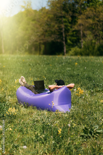 Handsome man lying on inflatable sofa lamzak working on laptop while resting on grass in park photo