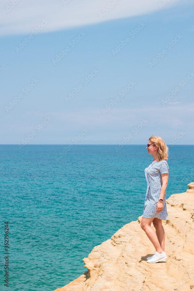 Young blonde caucasian woman tourist in dress and white sneakers standing on edge of sandy cliff rock above blue emerald crystal clear sea in summer sunny day.Vacation,travel,cruise concept Young blonde caucasian woman tourist in dress and white sneakers standing on edge of sandy cliff rock above blue emerald crystal clear sea in summer sunny day.Vacation,travel,cruise concept