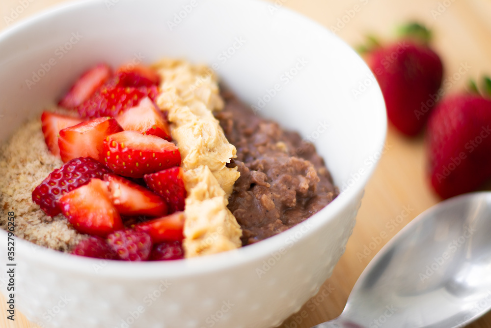 Chocolate cocoa oatmeal with almond meal, chopped strawberries and creamy peanut butter on a wooden surface. Strawberry, almonds, napkin and spoon in the background Chocolate cocoa oatmeal with almond meal, chopped strawberries and creamy peanut butter on a wooden surface. Strawberry, almonds, napkin and spoon in the background