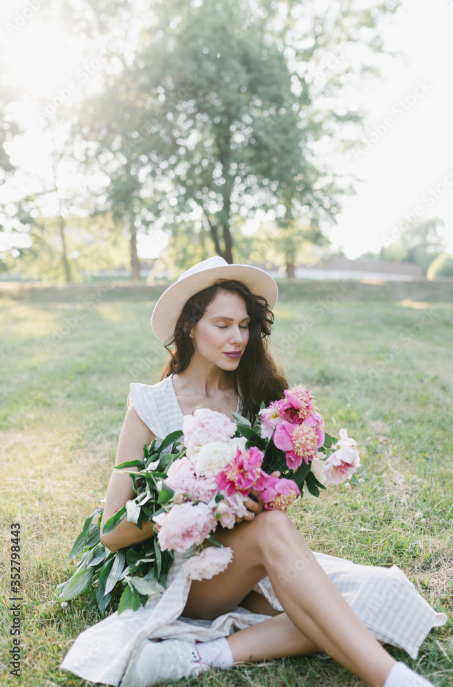 Fototapeta premium Portrait of a young woman in a hat and an armful of peonies. Beautiful stylish girl in a summer dress, a hat and a bouquet of flowers. Girl and flowers.