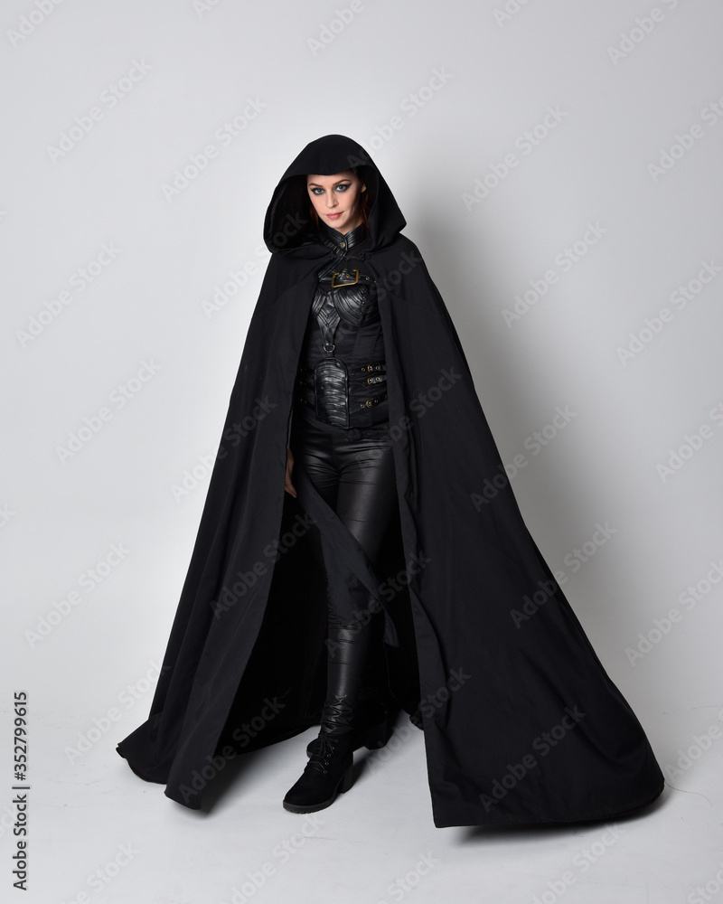 fantasy portrait of a woman with red hair wearing dark leather assassin costume with long black cloak. Full length standing pose isolated against a studio background. fantasy portrait of a woman with red hair wearing dark leather assassin costume with long black cloak. Full length standing pose isolated against a studio background.