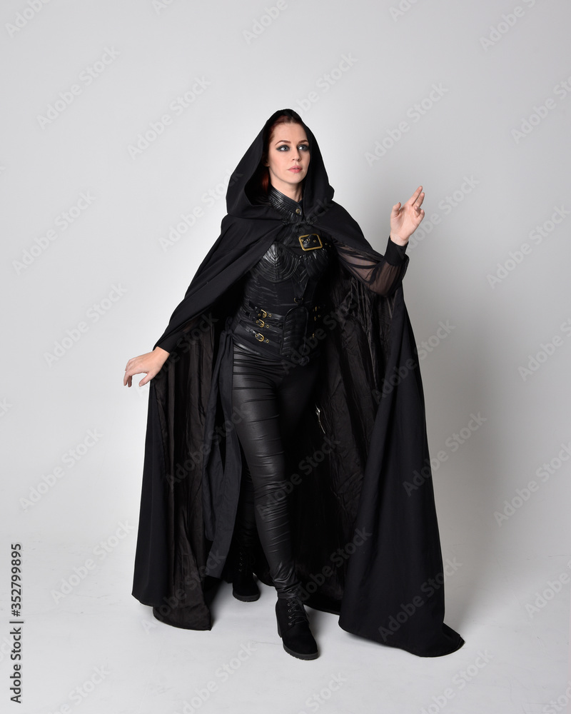 fantasy portrait of a woman with red hair wearing dark leather assassin costume with long black cloak. Full length standing pose  isolated against a studio background.