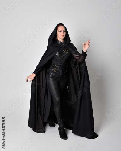 fantasy portrait of a woman with red hair wearing dark leather assassin costume with long black cloak. Full length standing pose  isolated against a studio background. © faestock
