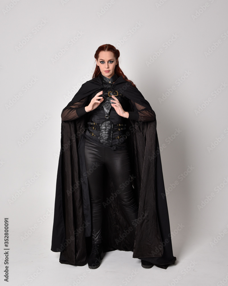 Fototapeta premium fantasy portrait of a woman with red hair wearing dark leather assassin costume with long black cloak. Full length standing pose isolated against a studio background.