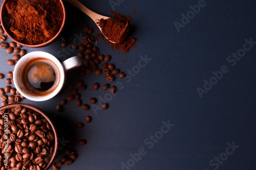 Fresh espresso cup, coffee beans and ground powder in clay bowls, wooden spoon on dark blue table surface. Closeup, top view, copy space. Coffee shop, morning, baristas workplace concept