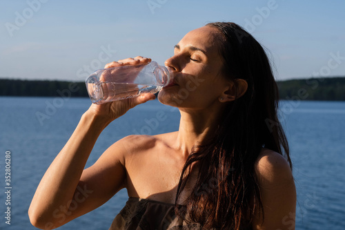 A beautiful woman in a bathing suit drinks fresh cool water from a bottle escaping the heat on the lake