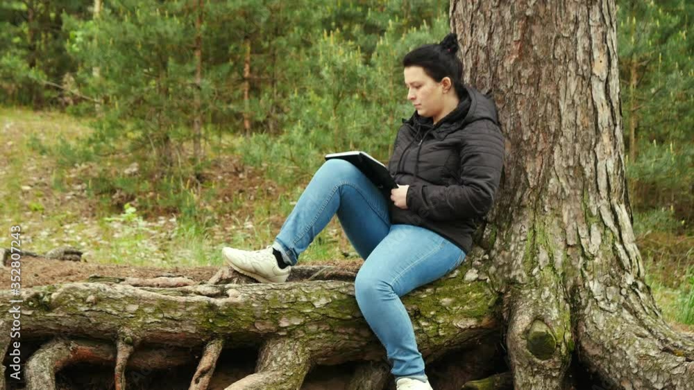 Girl sitting on large tree roots and writing something in a notebook. Girl sitting on large tree roots and writing something in a notebook.