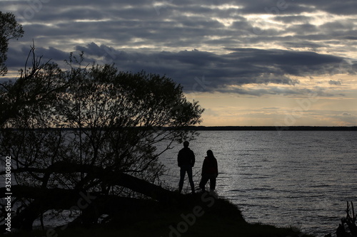 The silhouette of a guy and a girl on the shore of a lake with silvery water under a lighted sky with gray Cumulus clouds at dusk.A couple in love admires the beautiful landscape at sunset.Russia