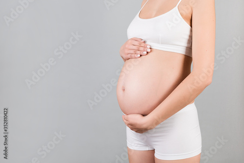 Cropped image of pregnant woman hugging her abdomen. Mother is wearing white underwear at gray background. Future parent is expecting her child. Copy space