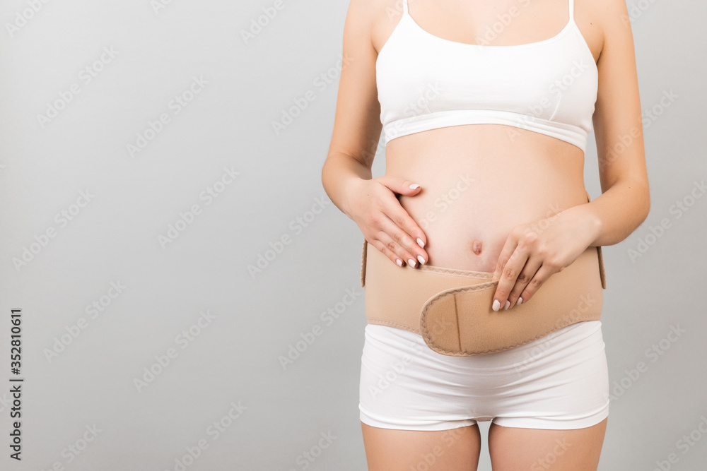 Close up of young pregnant woman in underwear dressing bandage on her belly at gray background with copy space. Orthopedic abdominal support belt concept