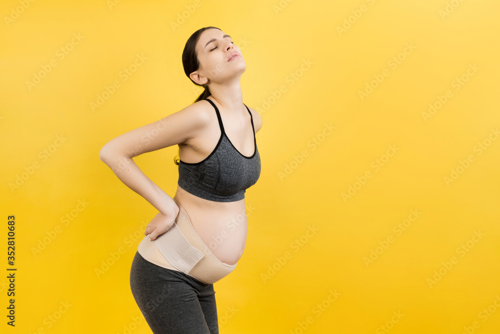 Portrait of supporting bandage against backache on pregnant woman at yellow background with copy space. Mother is suffering from pain in the back. Orthopedic abdominal support belt concept Portrait of supporting bandage against backache on pregnant woman at yellow background with copy space. Mother is suffering from pain in the back. Orthopedic abdominal support belt concept