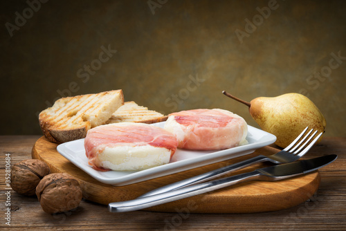 Grilled tomino cheese with speck (tomino boscaiolo) typical Italian Piedmontese cheese, with bread, walnut and pear on wooden table, copy space.
