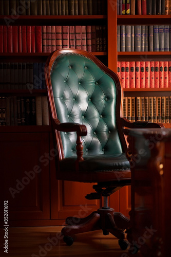 Dark solid interior of old library with books on the wooden shelves. Vintage classic chair green leather and red wood, antique table, luxury design with expensive natural details. Indoor, low exposure