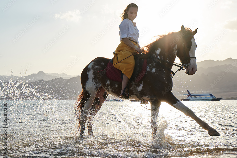 Caucasian blonde girl riding a horse through the water at sunrise Caucasian blonde girl riding a horse through the water at sunrise