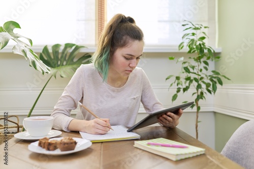 Teenager girl studying at home, student sitting at table using digital tablet