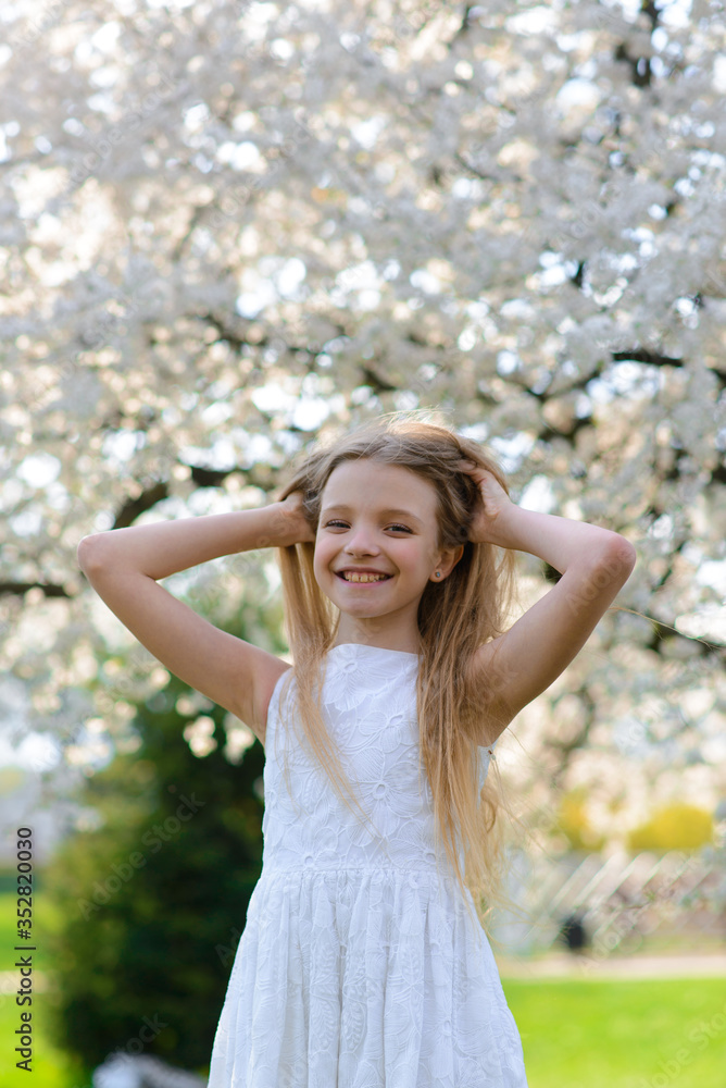 Beautiful blue-eyed girl with long blond hair in a white dress walking in the flower garden. summer bright, emotional photo.