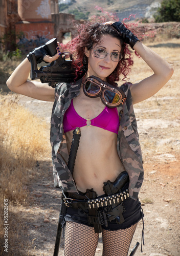 Very thin girl with pink hair and glasses dressed in military clothes carrying guns  machine guns, swords and other accessories for battle in a abandoned place