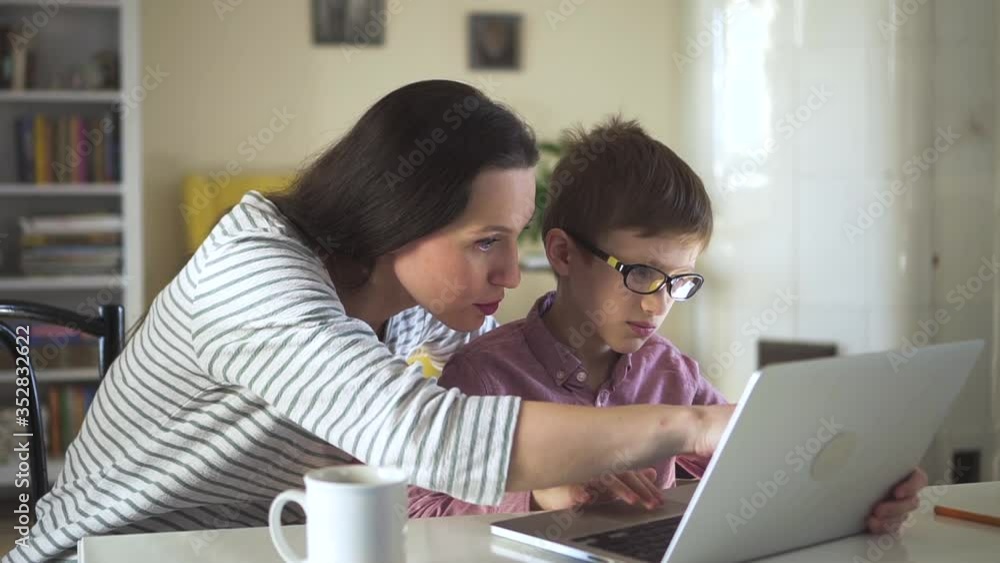 Young family of mom and kid using laptop at table in apartment during self-isolation spbd. European woman and child look at computer screen and talk with happy smiles while sitting at desk in light