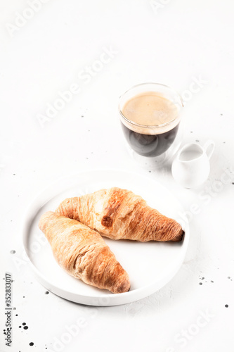 Two croissants on a plate next to a cup of coffee on light gray background.