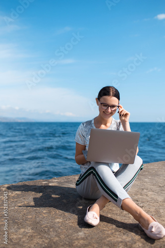 Beautiful young woman sitting with a laptop by the sea. Webinar online teaching concept. Online work.
