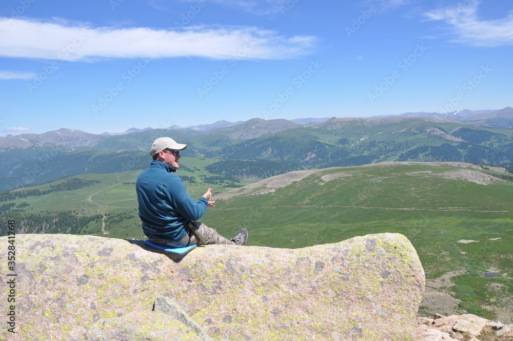 Naklejka premium A man in a sweatshirt and a baseball cap sits on a stone covered with lichen over a mountain valley