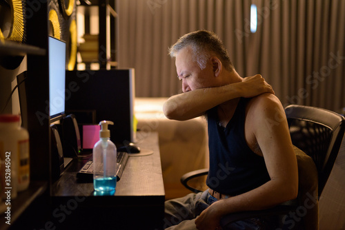 Stressed mature Japanese man covering cough with elbow while working from home