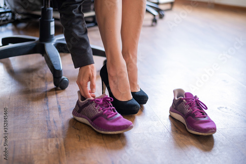 A business woman exchanges high heels for comfortable shoes in the workplace. A close-up of female hands takes off her black shoes and puts on colored sneakers in the office after a long working day.