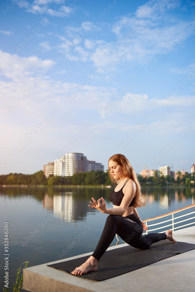Young woman doing yoga near the water, leading healthy lifestyle in nature, time to relax in the morning at the sunrise, meditation on the mat, copy space