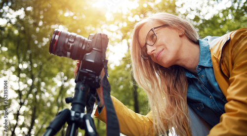 Low angle of blond woman looking at digital camera display taking pictures in green leafy forest photo