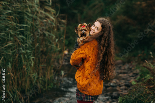 A teenage girl with long curly hair hugs a small dog with a bow on its head. Girl in an orange sweater in the open air
