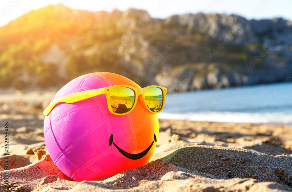 Rainbow colorful smiling beach ball dressed sunglasses on a sandy beach by tsea. Summer holidays concept.
