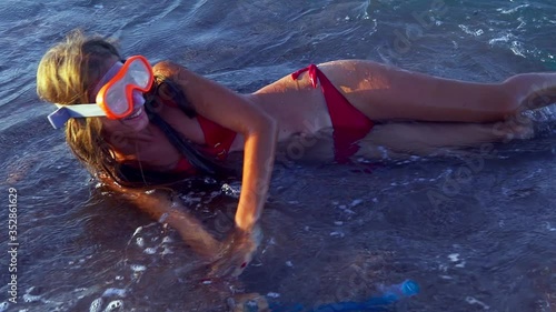 Beach vacation of girl sunburning by sea foam wave. Woman wearing red bikini and goggles with black diving fins turns from side to side along beach sand. Slow motion photo