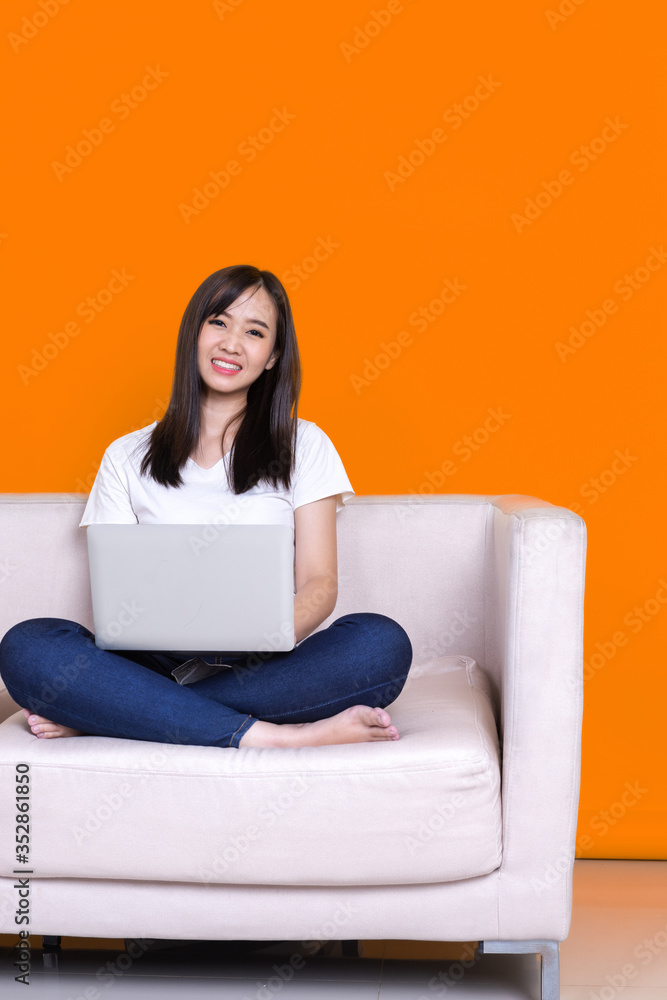 Asian woman smiling and working from home use laptop computer while sitting on sofa over isolate yellow background Asian woman smiling and working from home use laptop computer while sitting on sofa over isolate yellow background