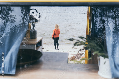 Young blond woman standing next to camper van overlooking the frozen winter sea. View through the open door with double exposure effect on window reflection.