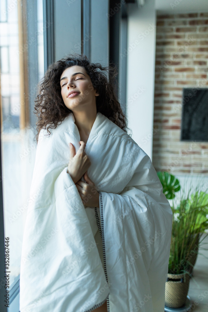 Young woman covered with blanket duver in the morning standing near window. Wake up in the cold morning Young woman covered with blanket duver in the morning standing near window. Wake up in the cold morning